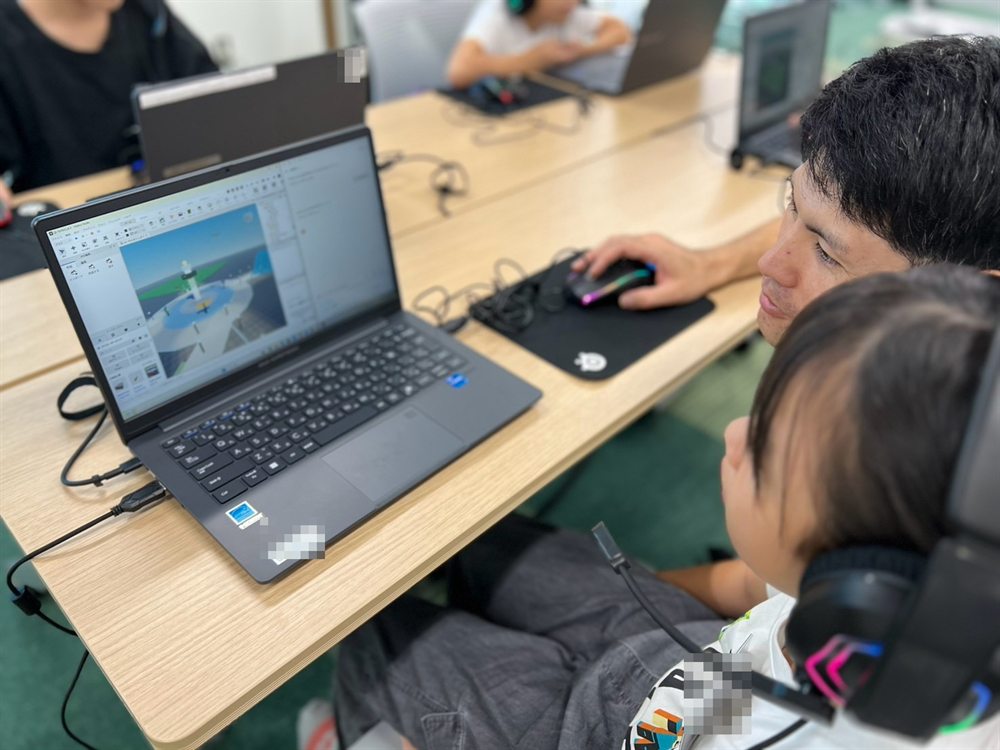 A teacher sits beside a child, looking together at the town on the screen