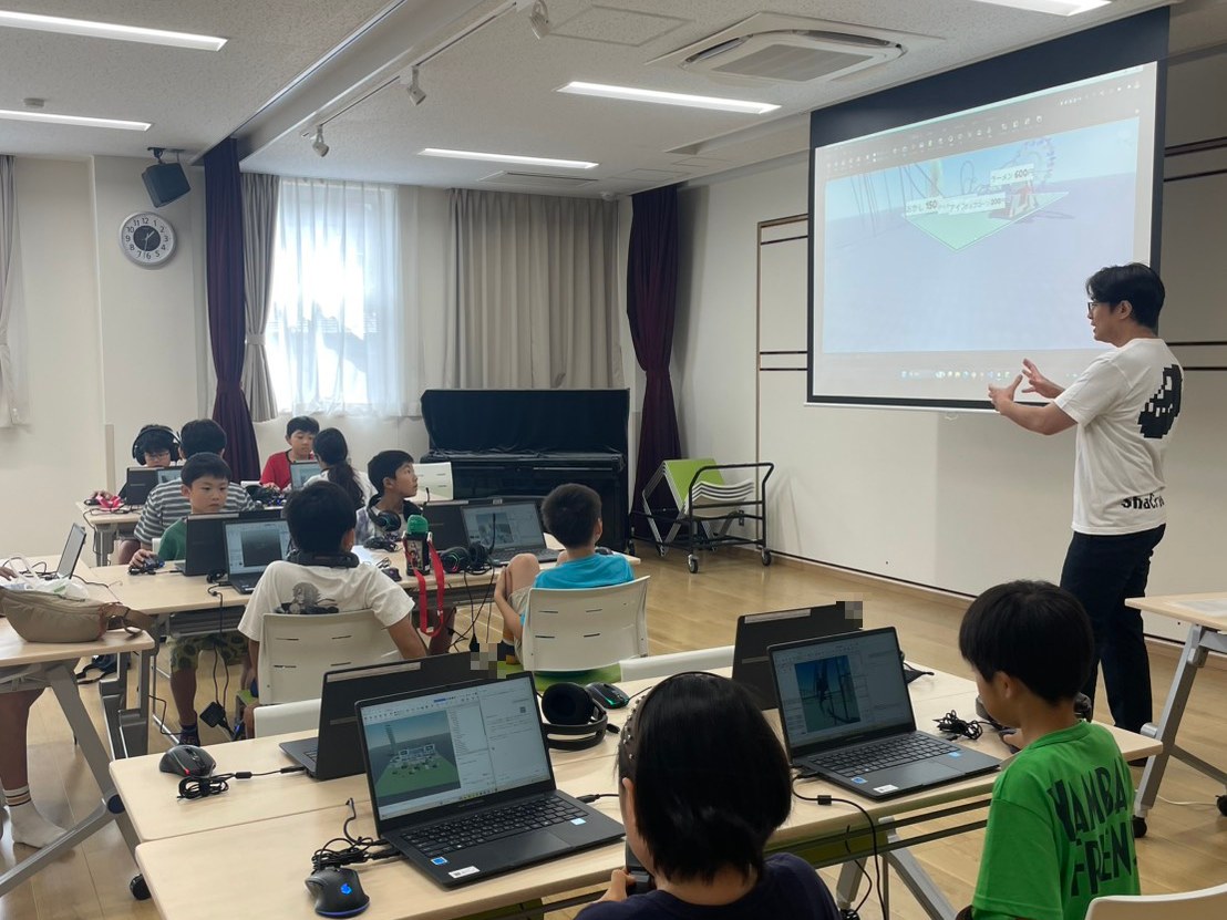 A teacher stands at the front of the room, speaking to all the children at their laptops